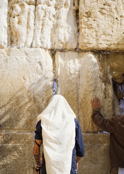 Two people are seen standing in front of a large, textured stone wall. One individual with a head covering appears to be in prayer, while the other has a hand placed on the wall. The wall has numerous pieces of paper inserted into its crevices.