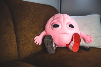 A soft, pink plush toy with a brain-like structure and a face with large eyes is sitting on a brown couch. The toy is wearing miniature sneakers, one red and one black, and there is a TV remote control lying next to it. The background includes a light-colored pillow.