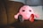 A soft, pink plush toy with a brain-like structure and a face with large eyes is sitting on a brown couch. The toy is wearing miniature sneakers, one red and one black, and there is a TV remote control lying next to it. The background includes a light-colored pillow.