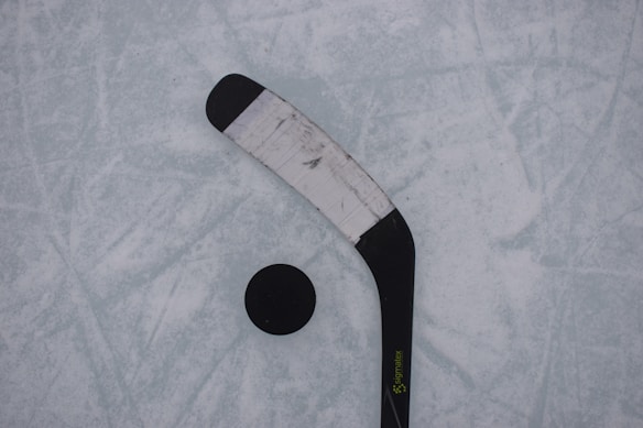 A close-up view of a hockey stick and puck resting on an ice rink. The hockey stick has a black blade with white tape wrapped around it. The puck is black and circular, positioned near the blade.