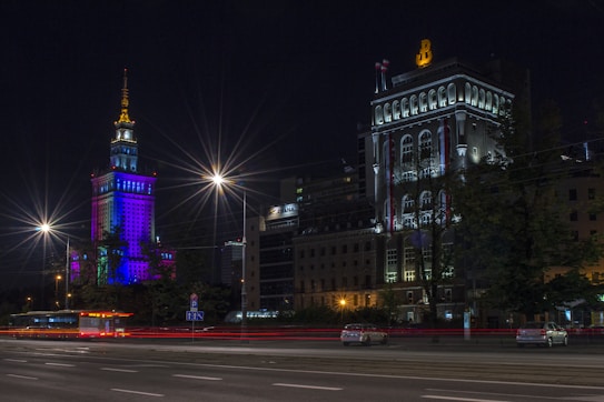 A vibrant cityscape at night with a skyscraper illuminated in colorful lights, including pink, blue, and purple hues. The street is lined with bright streetlamps emitting starburst effects against the dark sky. A bus stop shelter is visible on the side of the road, alongside a few cars with streaks of light from long exposure photography.