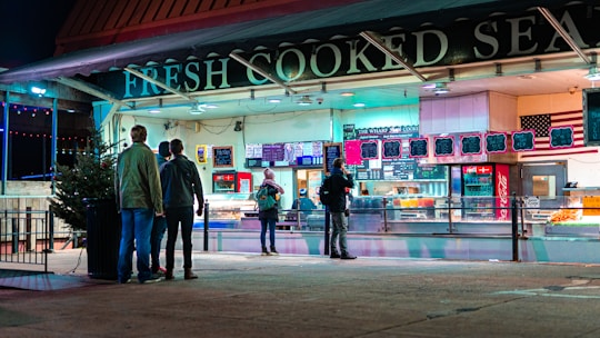 A group of people stands outside a brightly lit seafood restaurant at night. The sign above the entrance reads 'FRESH COOKED SEAFOOD.' The scene is vibrant with neon lights reflecting off the pavement, and there are colorful menus displayed above the counter.