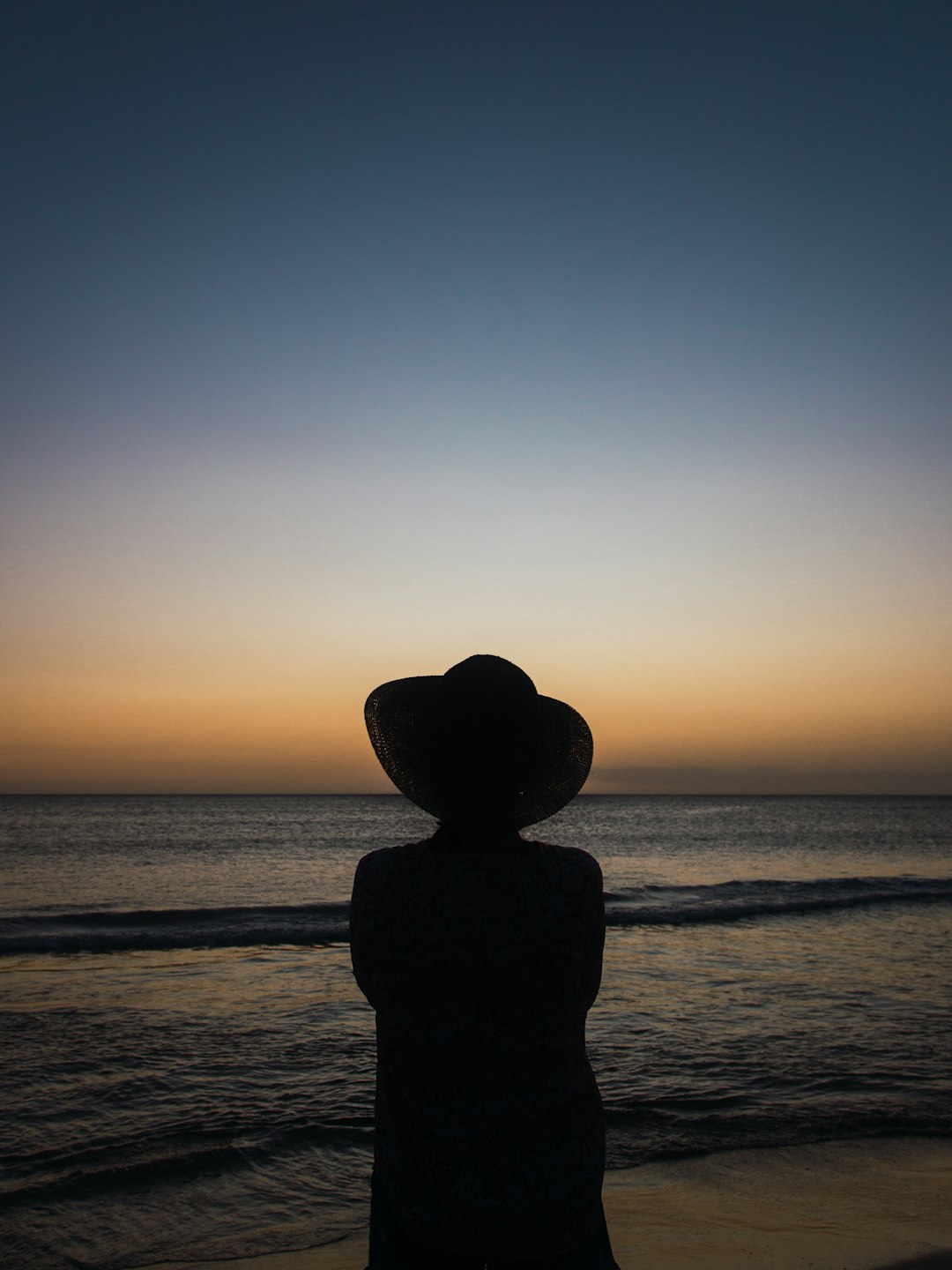 silhouette of person wearing hat standing on seashore during sunset