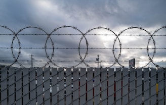 A barbed wire fence is silhouetted against a cloudy sky. The fence has large circular coils and vertical metal slats. In the background, some utility poles and a partially visible sign can be seen, adding to the industrial feel of the setting.