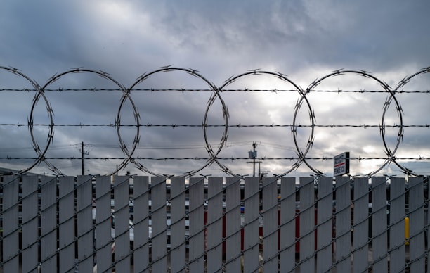 A barbed wire fence is silhouetted against a cloudy sky. The fence has large circular coils and vertical metal slats. In the background, some utility poles and a partially visible sign can be seen, adding to the industrial feel of the setting.