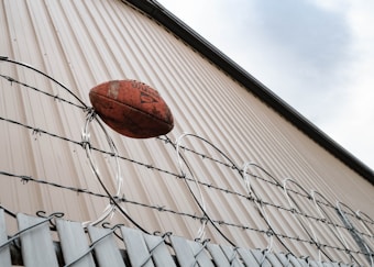 A football rests atop a barbed wire fence, positioned against the backdrop of a corrugated metal wall reaching up to a partly cloudy sky.