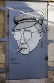A stylized black and white mural of a person wearing a hat and sunglasses, smoking a cigar, painted on a metal door. The artwork is signed with the text 'SHETACH TAL # JAFFADOORS' in the bottom left corner. The background of the door is painted blue and framed by a stone wall.