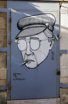 A stylized black and white mural of a person wearing a hat and sunglasses, smoking a cigar, painted on a metal door. The artwork is signed with the text 'SHETACH TAL # JAFFADOORS' in the bottom left corner. The background of the door is painted blue and framed by a stone wall.