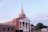 A large red-brick church with white columns and a tall steeple is set against a backdrop of a clear sky. The church features a classic architecture style with multiple arched windows and neatly trimmed bushes in the front.