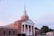 A large red-brick church with white columns and a tall steeple is set against a backdrop of a clear sky. The church features a classic architecture style with multiple arched windows and neatly trimmed bushes in the front.