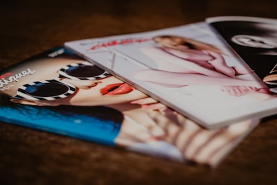 Three glossy fashion magazines are spread out on a surface. The cover of the left magazine features a close-up of a person wearing striped sunglasses and red lipstick. The center magazine shows a blurred image of a person in light-colored clothing, while the right magazine is partially visible and monochromatic.