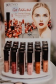 A display stand for Emani Vegan Cosmetics featuring Lip Shine Gloss with high impact colors. The poster includes an image of a woman with red lipstick and pomegranate seeds in the background. Below the poster, various shades of lip gloss are arranged in rows on a display.