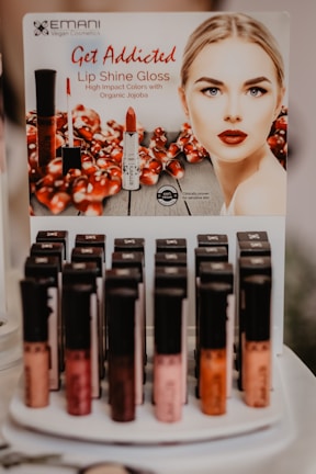 A display stand for Emani Vegan Cosmetics featuring Lip Shine Gloss with high impact colors. The poster includes an image of a woman with red lipstick and pomegranate seeds in the background. Below the poster, various shades of lip gloss are arranged in rows on a display.
