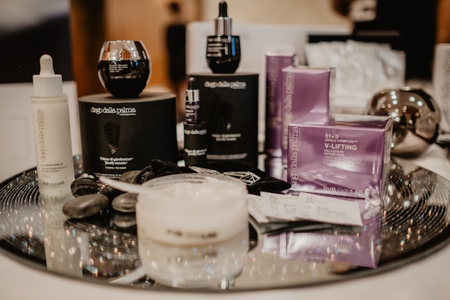 A collection of skincare products is arranged on a shiny surface, including bottles, jars, and boxes. The products feature black and purple packaging with visible branding. Some items have droppers, while another is a cream jar. Pebbles are decoratively placed beside the products.