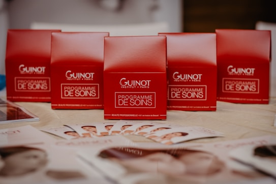 Several red boxes with 'Guinot Institut Paris' and 'Programme de Soins' written on them are arranged on a table. In the foreground, there are brochures displaying images of faces spread out on the table.