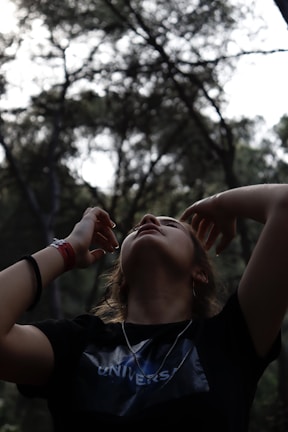 A person is standing in a forested area with their head tilted back, arms raised, and fingers spread near the face. The scene is framed by dense trees with muted light filtering through the branches. The individual wears a dark shirt with a visible text and has a relaxed, contemplative posture.