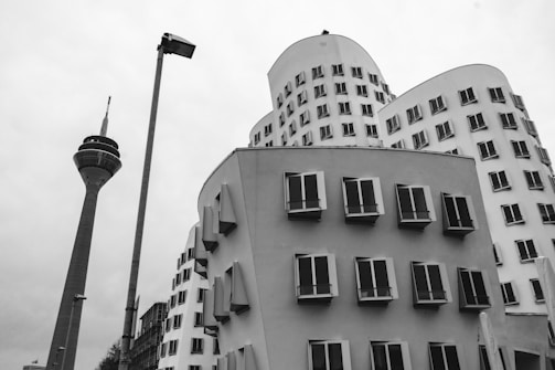 A black and white photograph showing modern architectural buildings with curved designs and numerous small windows. A tall communications tower stands nearby, alongside a streetlight pole.