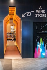 A uniquely designed entrance in the shape of a wine bottle leads to a liquor store, with shelves of wine bottles visible inside. The exterior is dark with bright, warm lighting accentuating the entrance, and there are colorful geometric designs on an adjacent wall.