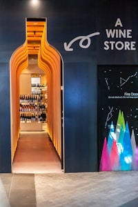 A uniquely designed entrance in the shape of a wine bottle leads to a liquor store, with shelves of wine bottles visible inside. The exterior is dark with bright, warm lighting accentuating the entrance, and there are colorful geometric designs on an adjacent wall.