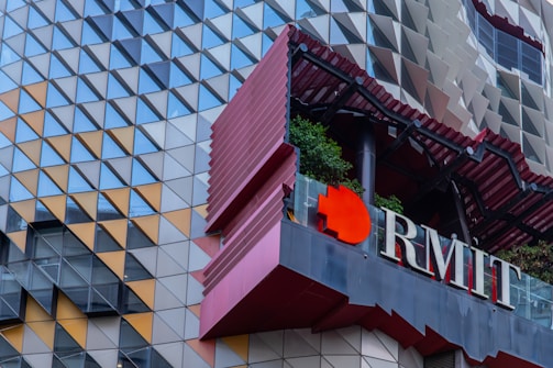 An architectural facade composed of a geometric pattern made up of triangular panels. The panels are arranged in a repeated sequence with shades of gray, orange, and blue. The structure features a prominent red accent and vegetation, complemented by the letters 'RMIT' on the building.
