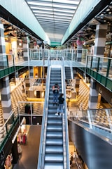 A modern shopping mall with multiple levels, featuring a large central escalator flanked by glass railings. The architecture includes geometric patterns on the floors and a high ceiling with skylights that allow natural light to flood the space. Various stores and small kiosks line the walkways, and there are decorative elements such as hanging plants and colorful flowers. Shoppers are visible browsing and walking on different levels.