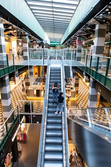 A modern shopping mall with multiple levels, featuring a large central escalator flanked by glass railings. The architecture includes geometric patterns on the floors and a high ceiling with skylights that allow natural light to flood the space. Various stores and small kiosks line the walkways, and there are decorative elements such as hanging plants and colorful flowers. Shoppers are visible browsing and walking on different levels.