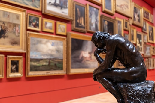 A detailed view of an art gallery, featuring numerous framed paintings displayed on a red wall. In the foreground, a prominent bronze sculpture of a thinker in a contemplative pose is positioned on a stone base.