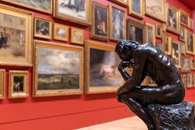 A detailed view of an art gallery, featuring numerous framed paintings displayed on a red wall. In the foreground, a prominent bronze sculpture of a thinker in a contemplative pose is positioned on a stone base.