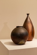 Two ceramic vessels are placed on a white pedestal against a neutral background. The vessels have a textured, handcrafted appearance with one being spherical and dark-colored, while the other is taller, teardrop-shaped with a warm, earthy hue.
