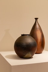 Two ceramic vessels are placed on a white pedestal against a neutral background. The vessels have a textured, handcrafted appearance with one being spherical and dark-colored, while the other is taller, teardrop-shaped with a warm, earthy hue.