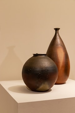 Two ceramic vessels are placed on a white pedestal against a neutral background. The vessels have a textured, handcrafted appearance with one being spherical and dark-colored, while the other is taller, teardrop-shaped with a warm, earthy hue.