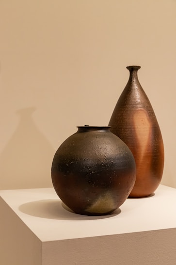 Two ceramic vessels are placed on a white pedestal against a neutral background. The vessels have a textured, handcrafted appearance with one being spherical and dark-colored, while the other is taller, teardrop-shaped with a warm, earthy hue.