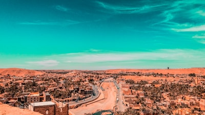 A panoramic view of a city with a road cutting through the center, surrounded by numerous palm trees and low-rise buildings. The sky is vibrantly colored with turquoise, contrasting with the warm tones of the earthly landscape.