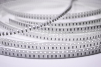 Spools of electronic components arranged in a coiled, layered pattern. The tape has a series of evenly spaced black rectangles with numbers, likely indicating each component. The plastic material has regularly spaced white circles along the edges.