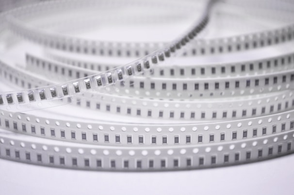 Spools of electronic components arranged in a coiled, layered pattern. The tape has a series of evenly spaced black rectangles with numbers, likely indicating each component. The plastic material has regularly spaced white circles along the edges.