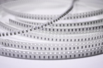 Spools of electronic components arranged in a coiled, layered pattern. The tape has a series of evenly spaced black rectangles with numbers, likely indicating each component. The plastic material has regularly spaced white circles along the edges.