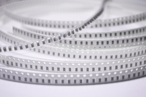 Spools of electronic components arranged in a coiled, layered pattern. The tape has a series of evenly spaced black rectangles with numbers, likely indicating each component. The plastic material has regularly spaced white circles along the edges.
