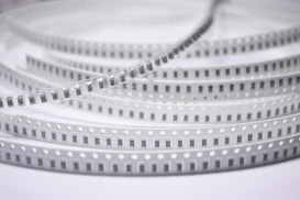 Spools of electronic components arranged in a coiled, layered pattern. The tape has a series of evenly spaced black rectangles with numbers, likely indicating each component. The plastic material has regularly spaced white circles along the edges.