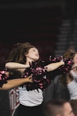 A female cheerleader energetically dances with pom-poms held high, captured in mid-motion with her hair flowing back. She is wearing a white top and black pants, performing in front of a blurred background that suggests a sports event.
