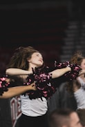 A female cheerleader energetically dances with pom-poms held high, captured in mid-motion with her hair flowing back. She is wearing a white top and black pants, performing in front of a blurred background that suggests a sports event.