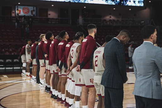 A group of basketball players and coaches stand in a line on the court, with hands behind their backs, possibly during a pre-game ritual or national anthem. They are wearing matching team outfits with numbers and names on their jerseys. The lighting in the arena is subdued with a focus on the court.