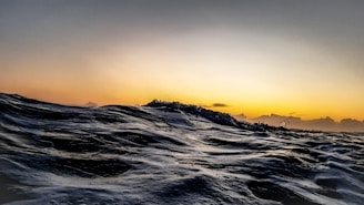 Waves dominate the foreground, with a dynamic, textured surface reflecting the ambient light. The horizon showcases a transition from deep sea to the warm, glowing colors of a sunset sky, where hints of cloud formations are silhouetted.