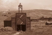 A sepia-toned photograph depicting a rustic, abandoned building with wooden planks and structures. In the background, several old, deserted wooden houses are situated on a barren landscape with sparse vegetation and rolling hills.