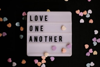 A lightbox with the message 'LOVE ONE ANOTHER' in block letters is surrounded by small, colorful heart-shaped candies scattered on a dark background.
