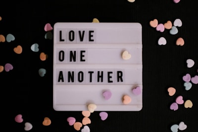 A lightbox with the message 'LOVE ONE ANOTHER' in block letters is surrounded by small, colorful heart-shaped candies scattered on a dark background.