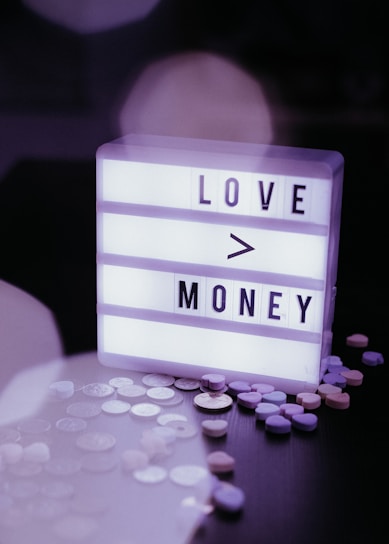 A lightbox with the text 'LOVE > MONEY' surrounded by scattered coins and colorful heart-shaped candies on a dark surface.