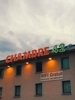 A building facade featuring large neon letters spelling 'CHAMBRE 42€' with a sign offering free WiFi in all buildings. The signage is prominently illuminated, contrasting against a cloudy evening sky.