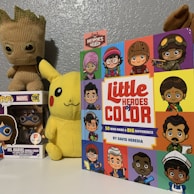 A children's book titled 'Little Heroes of Color' featuring cartoon illustrations of diverse characters with different cultural backgrounds and attire is prominently displayed. Beside it, a plush toy of Pikachu with a cheerful expression and a pop figurine of Ms. Marvel in a clear box are visible. In the background, there is another character toy resembling Groot from the 'Guardians of the Galaxy'. The wall is painted a greyish-blue, creating a calm backdrop for the colorful items.