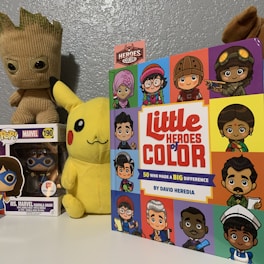 A children's book titled 'Little Heroes of Color' featuring cartoon illustrations of diverse characters with different cultural backgrounds and attire is prominently displayed. Beside it, a plush toy of Pikachu with a cheerful expression and a pop figurine of Ms. Marvel in a clear box are visible. In the background, there is another character toy resembling Groot from the 'Guardians of the Galaxy'. The wall is painted a greyish-blue, creating a calm backdrop for the colorful items.