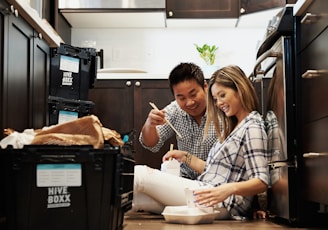 A couple is seated on the floor of a kitchen surrounded by packing boxes labeled 'Hive Boxx'. They are smiling and appear to be enjoying unpacking or eating together. The kitchen has dark wooden cabinets and a white countertop with a potted plant visible in the background.